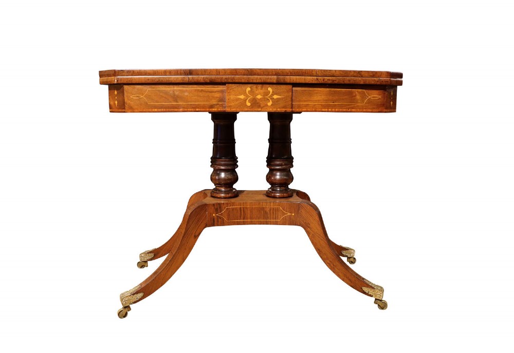 A Regency period rosewood foldover card table featuring marquetry inlay, twin turned pedestals, and four curved legs with brass sabots and castors.