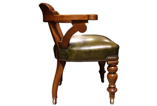 A Victorian golden oak library chair featuring a curved backrest, turned front legs with castors, and an olive green leather seat trimmed with brass studs. View V