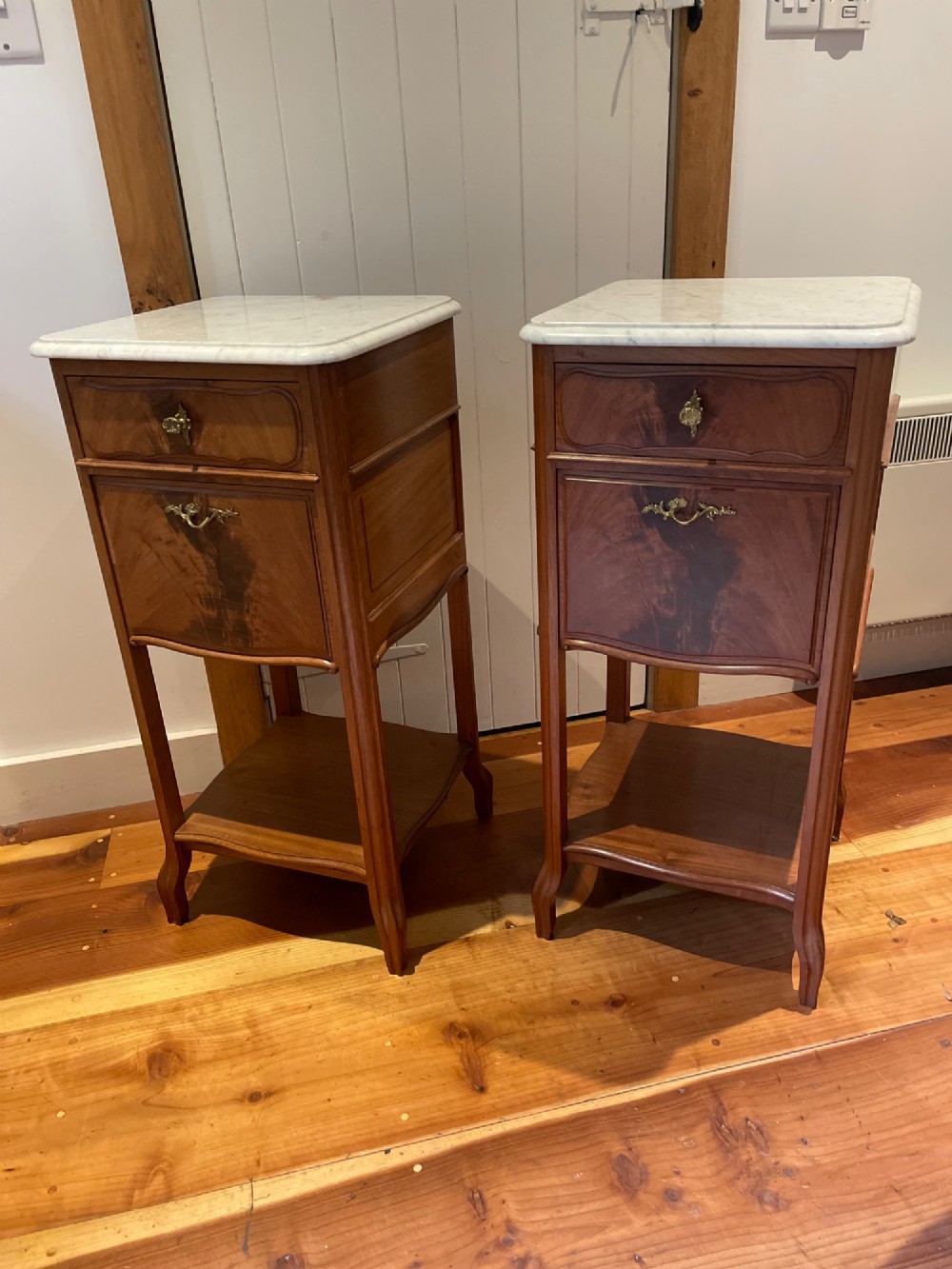 A pair of tall French wooden bedside cupboards featuring inset white marble tops, single drawers, cupboard doors, and lower open shelves.