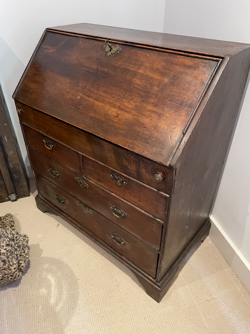 An 18th-century Queen Anne dark oak bureau featuring a slanted fall-front writing flap over drawers with brass drop handles.