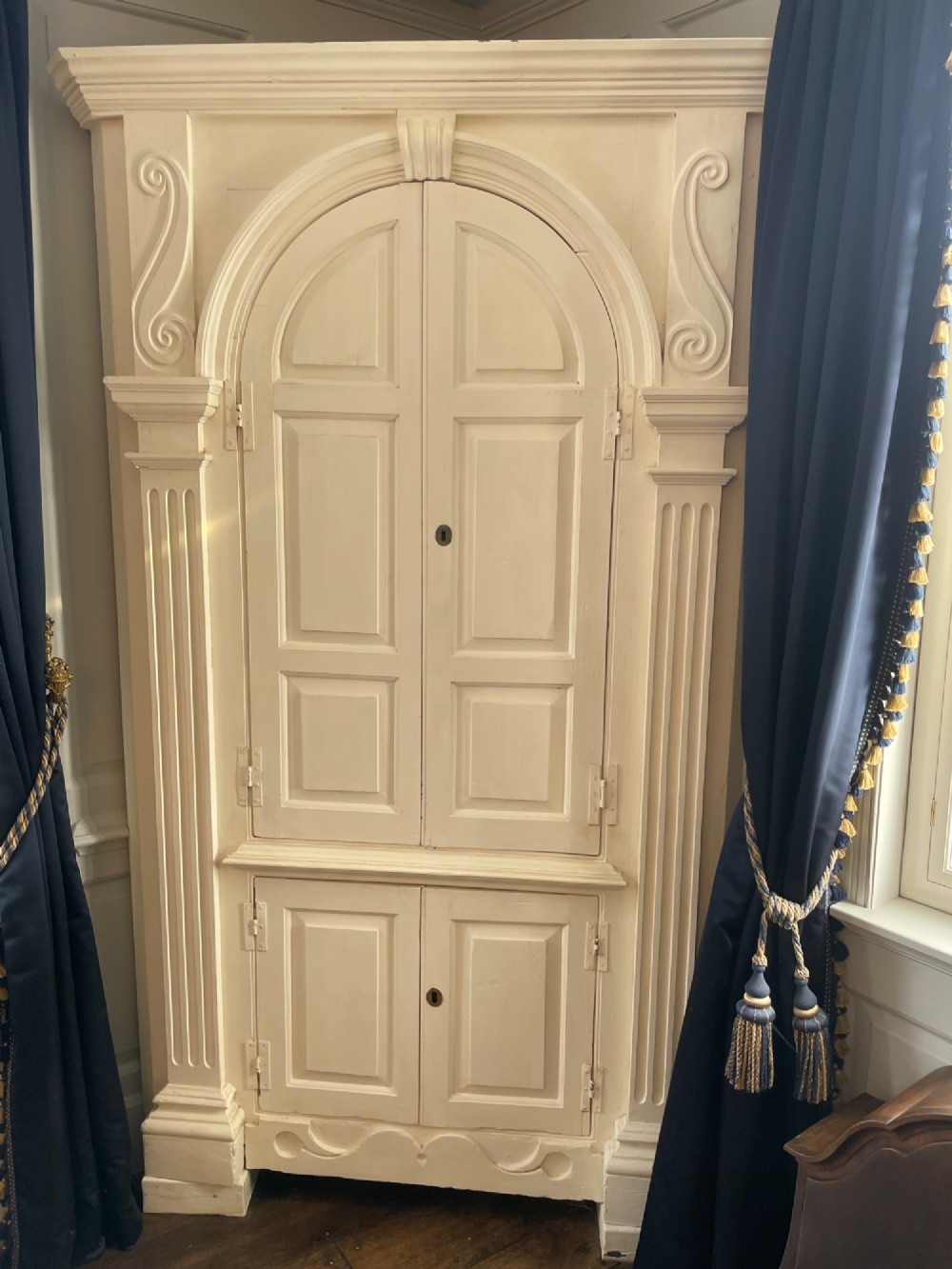 A tall cream-painted pine corner cupboard with open panel doors revealing an arched dark wood shelf interior and lower cabinet. View V