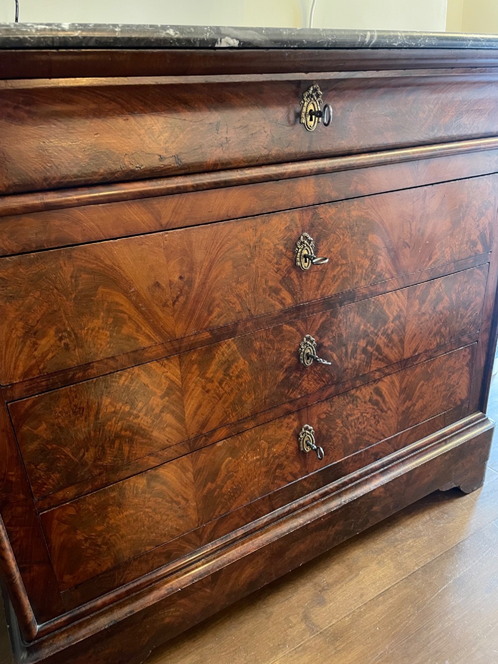 A French mahogany chest of drawers featuring a dark grey veined marble top, a pull-out brushing slide, three main drawers, and brass keyhole escutcheons. View III