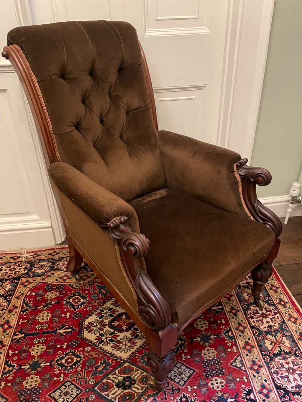 A Victorian mahogany library chair featuring dark brown velvet button-tufted upholstery, carved scroll arms, and brass castors.