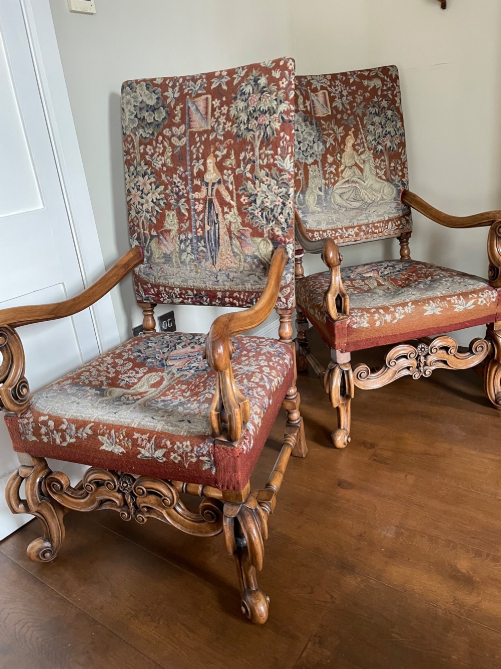 A pair of high-backed William and Mary style walnut armchairs with scroll arms, carved decorative stretchers, and pictorial woven tapestry upholstery.