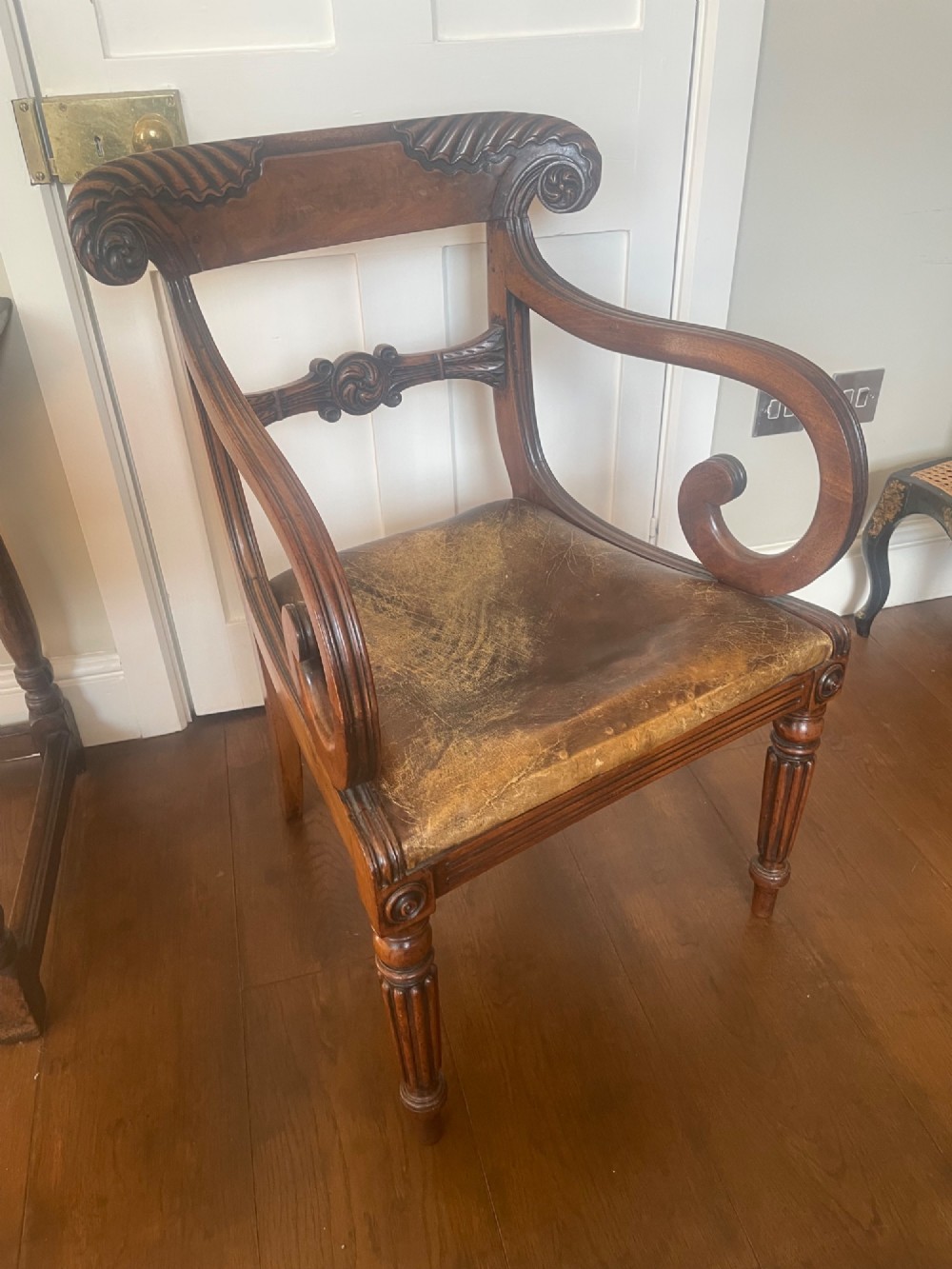 A William IV mahogany carver armchair featuring carved scroll arms, an upholstered distressed brown leather seat, decorative crest carvings, and turned fluted front legs.