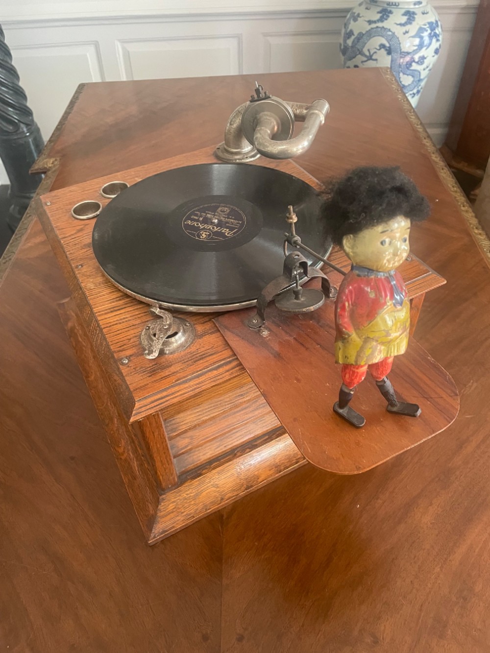 A square oak tabletop gramophone featuring a side winding crank, metal tonearm, black vinyl record, and a painted metal dancing figure attached to an extended wooden platform. View II
