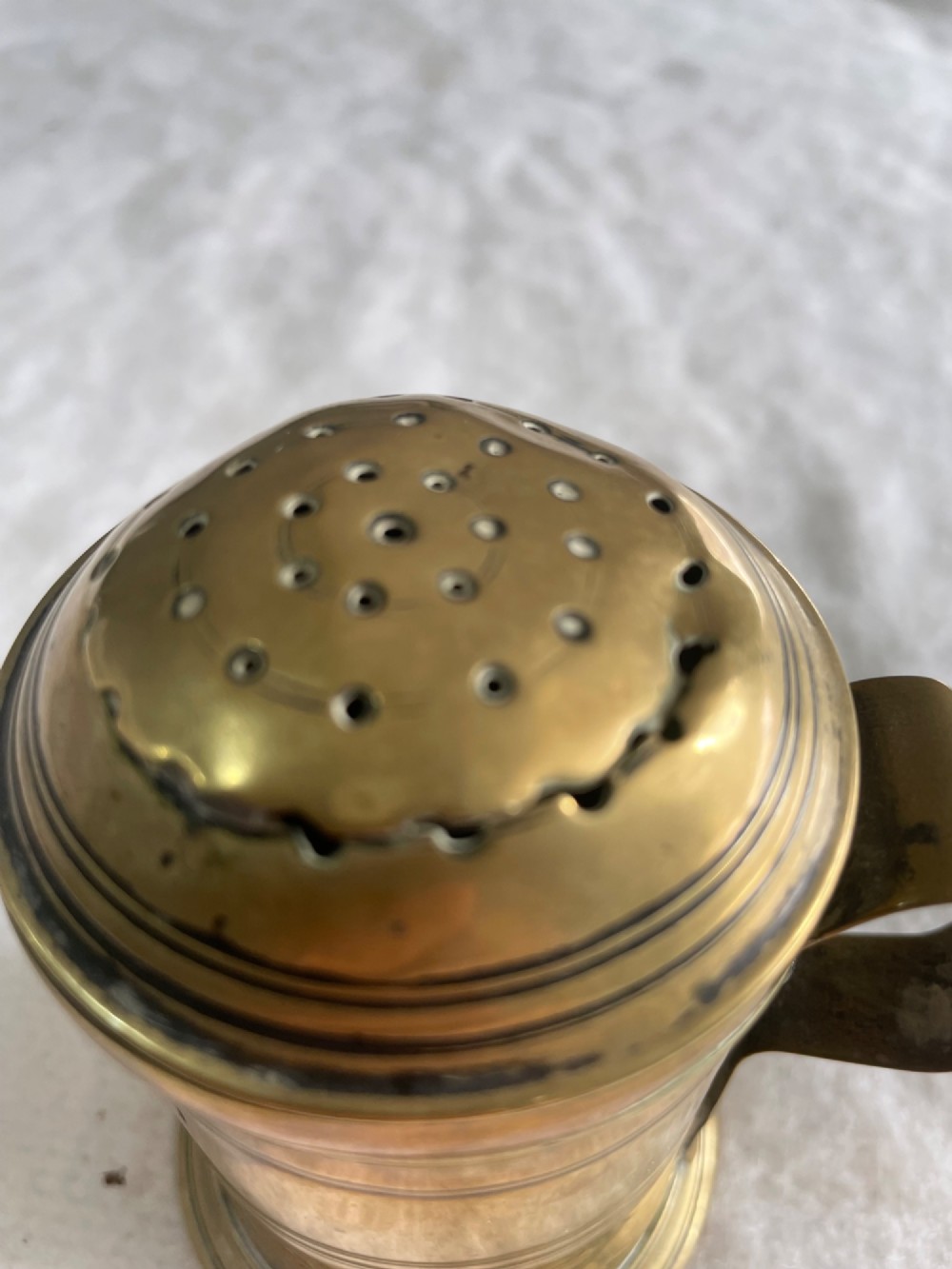 A small cylindrical antique brass ink sander with a perforated domed lid, side handle, and incised decorative ring details. View II