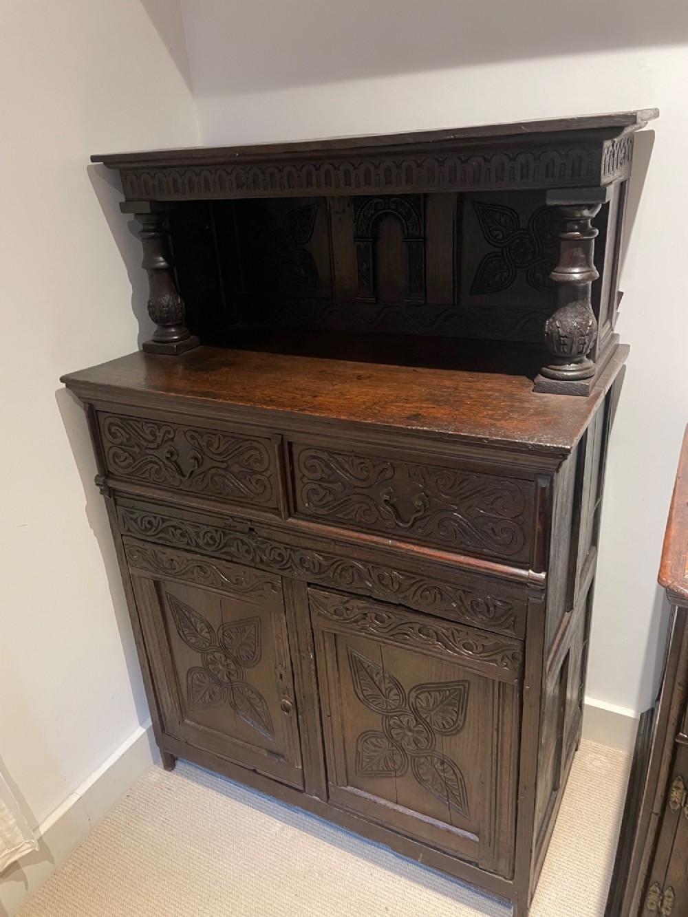 A dark carved oak court cupboard featuring an open upper shelf supported by turned balusters above two carved drawers and two panelled cupboard doors.