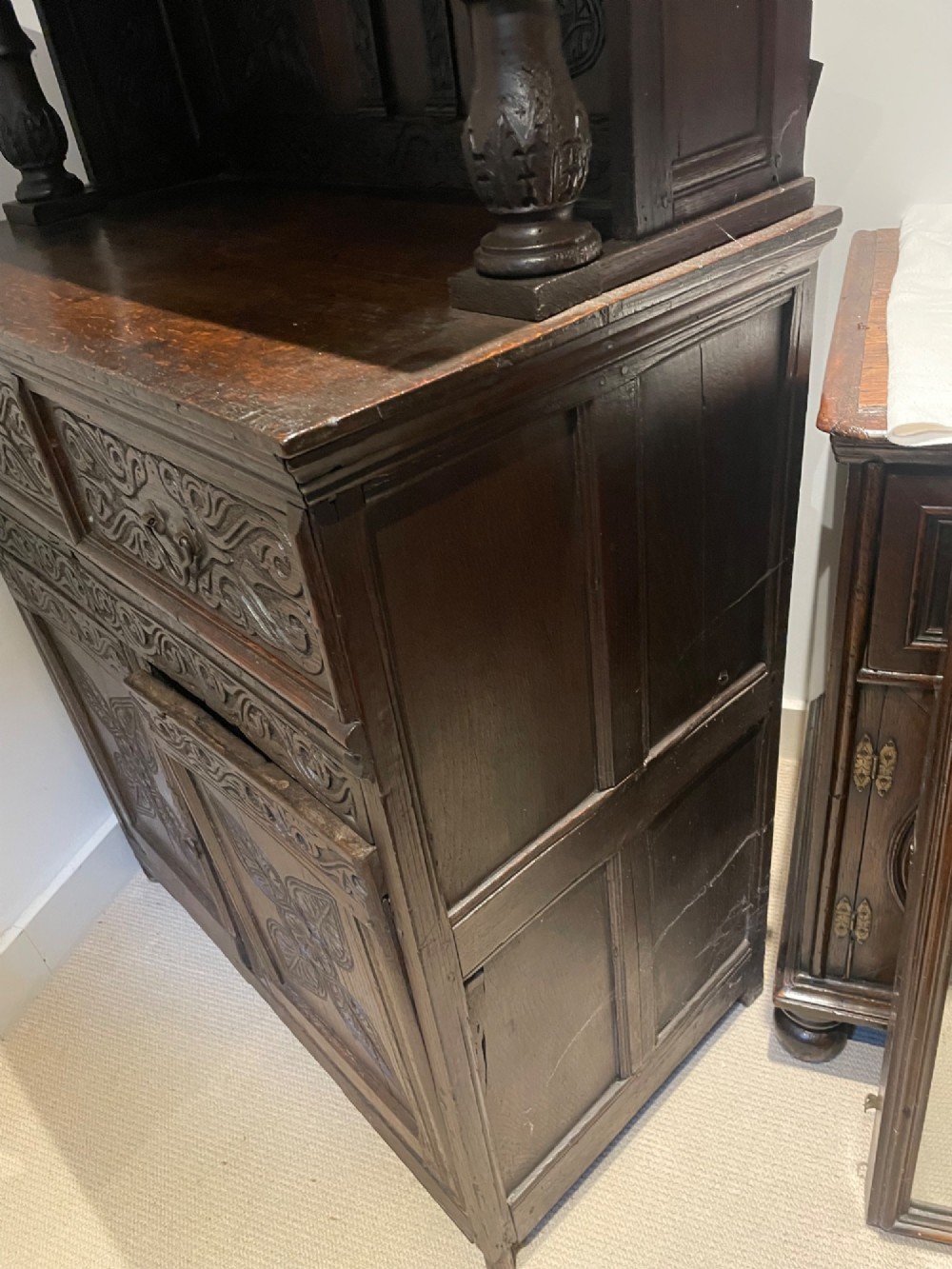 A dark carved oak court cupboard featuring an open upper shelf supported by turned balusters above two carved drawers and two panelled cupboard doors. View II