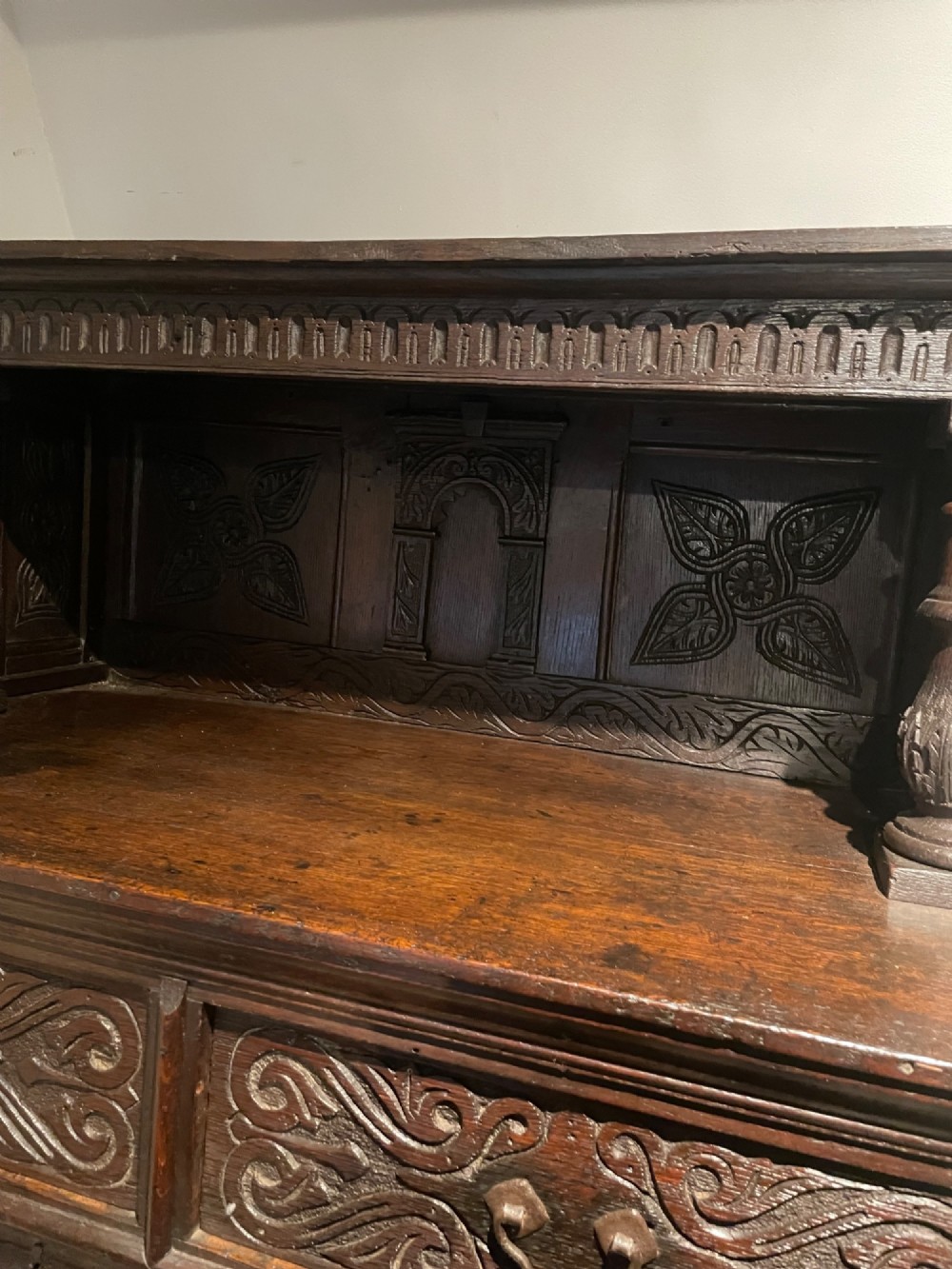 A dark carved oak court cupboard featuring an open upper shelf supported by turned balusters above two carved drawers and two panelled cupboard doors. View IV