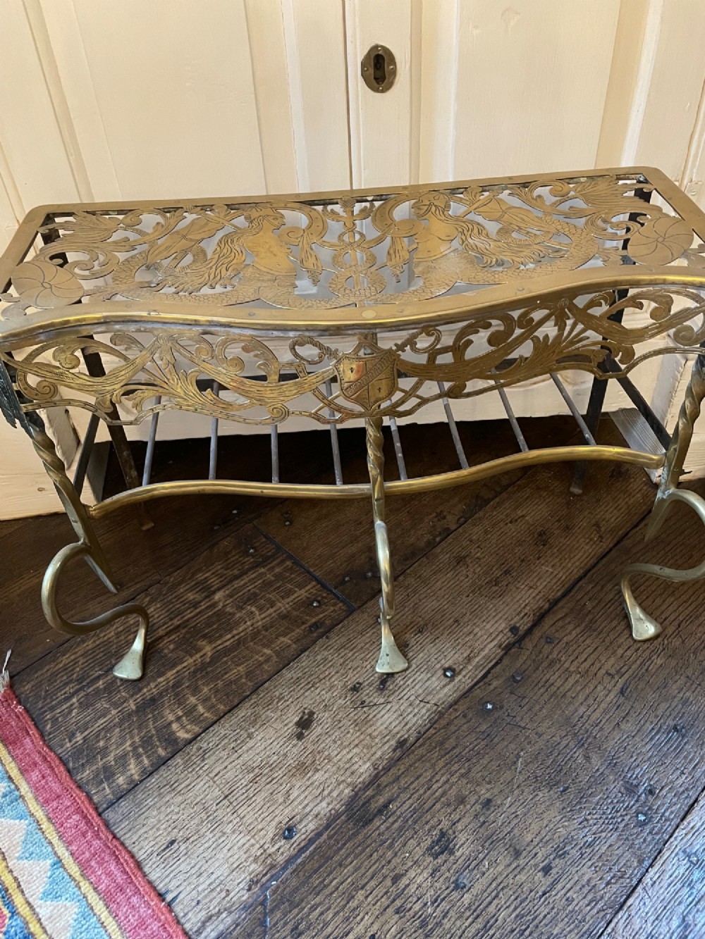 A two-tier brass fireplace fender featuring an etched mermaid top plate, a pierced fretwork skirt with a shield crest, and spiralled cabriole legs on a wooden floor.