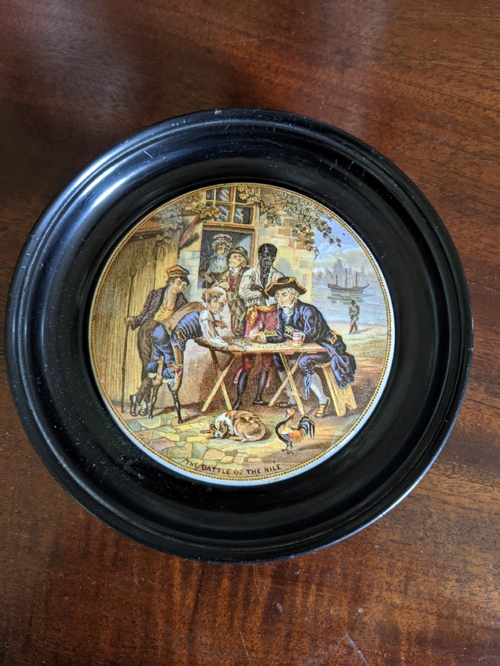 A circular Victorian Prattware ceramic pot lid titled The Battle of the Nile framed in a round ebonised wood frame.