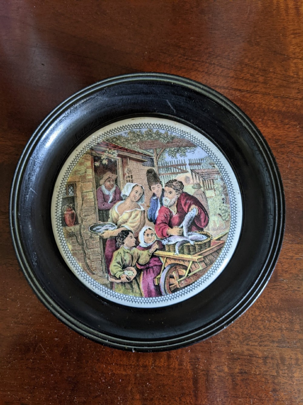 A circular Victorian polychrome ceramic pot lid depicting a historical fishmonger scene, set within a turned ebonised wood frame.