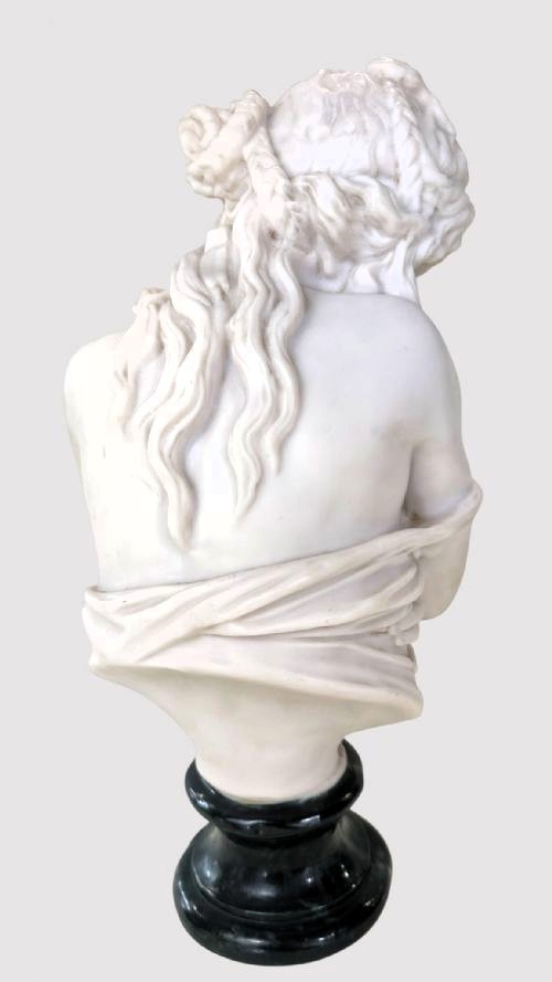 A carved white marble bust of a young woman with coiled hair and draped classical attire, mounted on a dark green marble circular plinth. View II