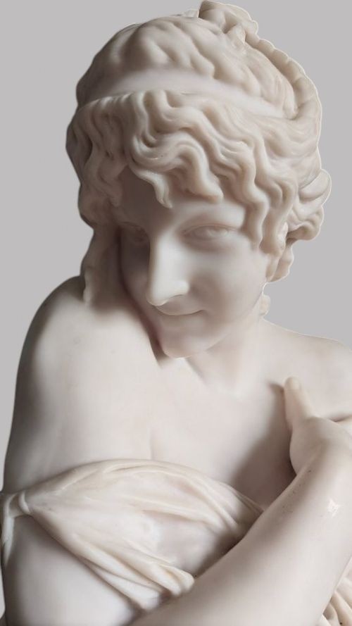 A carved white marble bust of a young woman with coiled hair and draped classical attire, mounted on a dark green marble circular plinth. View V