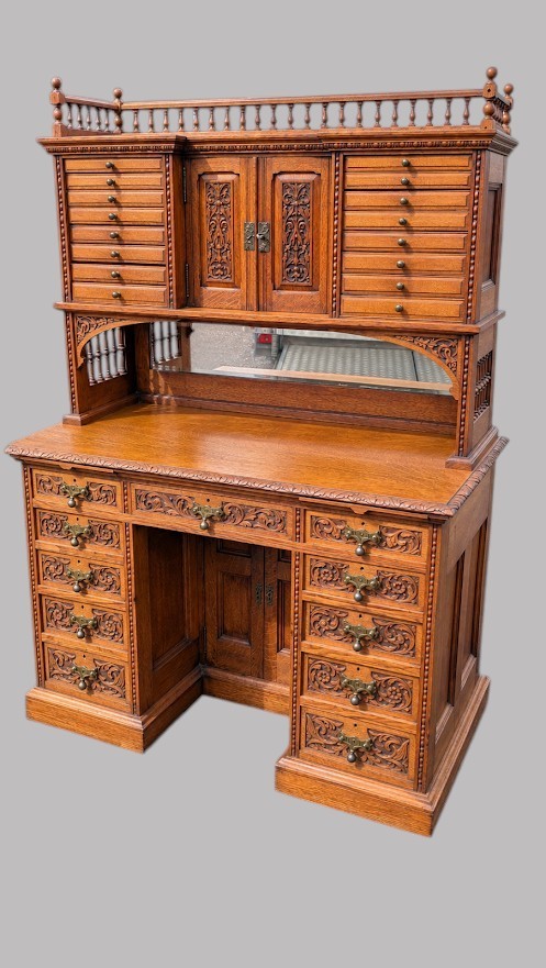 A late Victorian quarter-sawn oak collectors desk featuring carved pedestals, multiple shallow drawer tiers, a central cupboard, and a top spindle gallery. View V