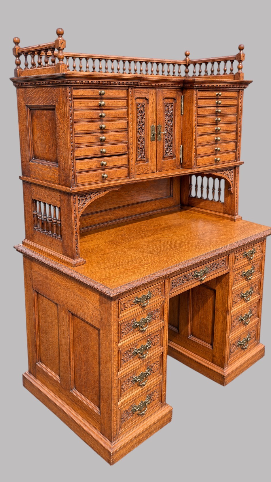 A late Victorian quarter-sawn oak collectors desk featuring carved pedestals, multiple shallow drawer tiers, a central cupboard, and a top spindle gallery. View VI