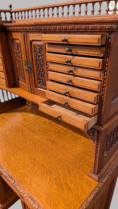 A late Victorian quarter-sawn oak collectors desk featuring carved pedestals, multiple shallow drawer tiers, a central cupboard, and a top spindle gallery. View VII