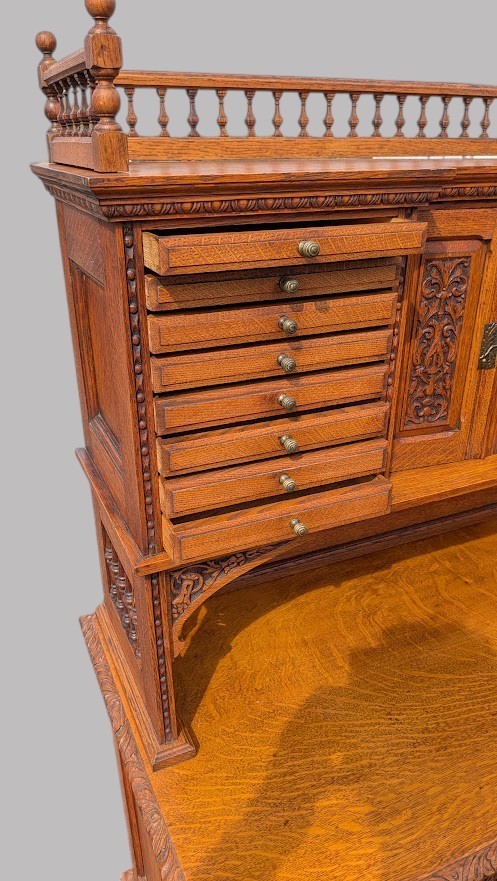 A late Victorian quarter-sawn oak collectors desk featuring carved pedestals, multiple shallow drawer tiers, a central cupboard, and a top spindle gallery. View VIII