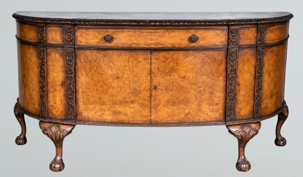 A bow-front burr walnut sideboard with carved pilasters, twin cupboard doors, a top drawer, and four carved cabriole legs ending in ball and claw feet.