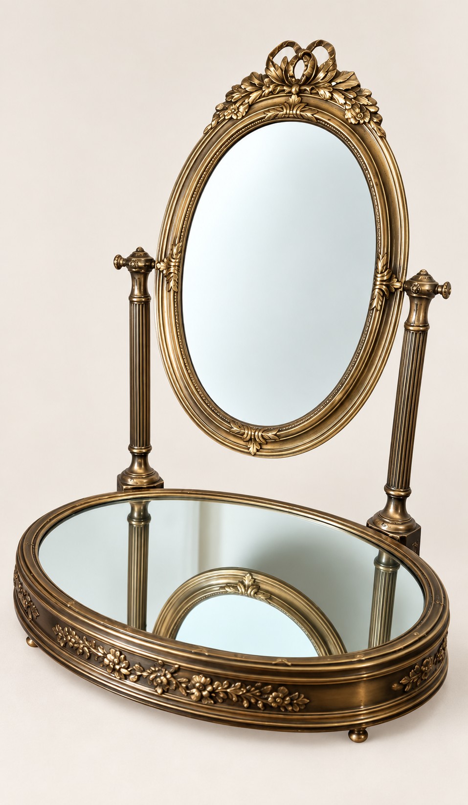 An antique late 19th-century French brass vanity mirror featuring an oval pivoting glass frame adorned with a bow crest, supported by two fluted columns resting on a circular mirrored base. View III