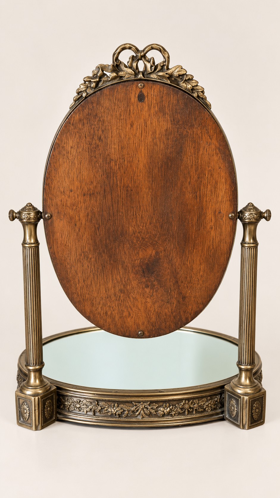 An antique late 19th-century French brass vanity mirror featuring an oval pivoting glass frame adorned with a bow crest, supported by two fluted columns resting on a circular mirrored base. View IV