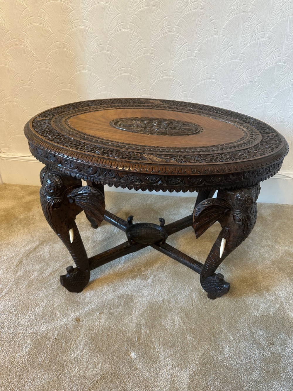 An oval dark brown padouk wood side table featuring hand-carved floral borders, a relief central panel, and four carved elephant head legs with white tusks.