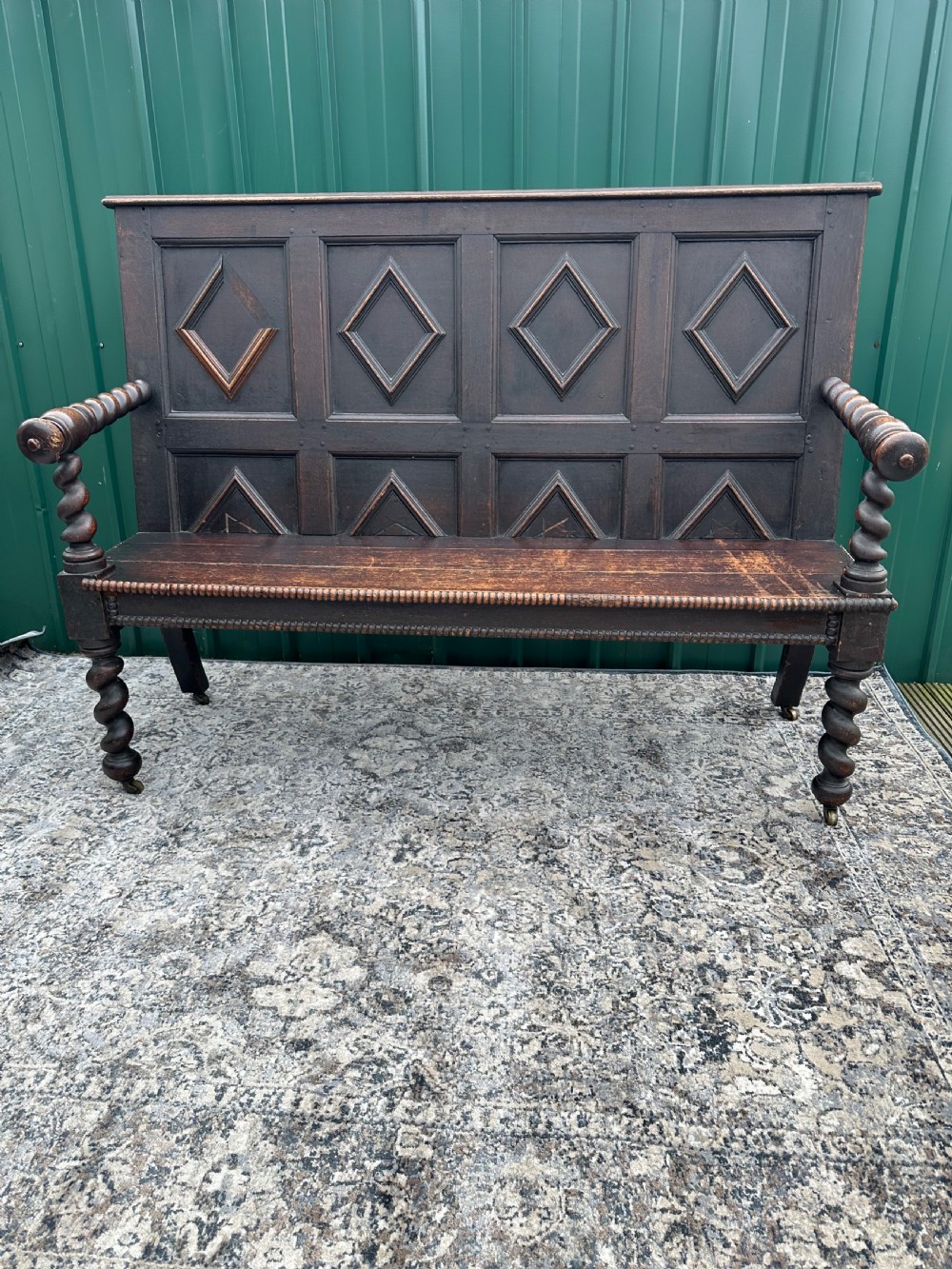 A dark oak 19th-century settle featuring a four-panel back with geometric diamond moulding, barley twist supports, and small brass castors.