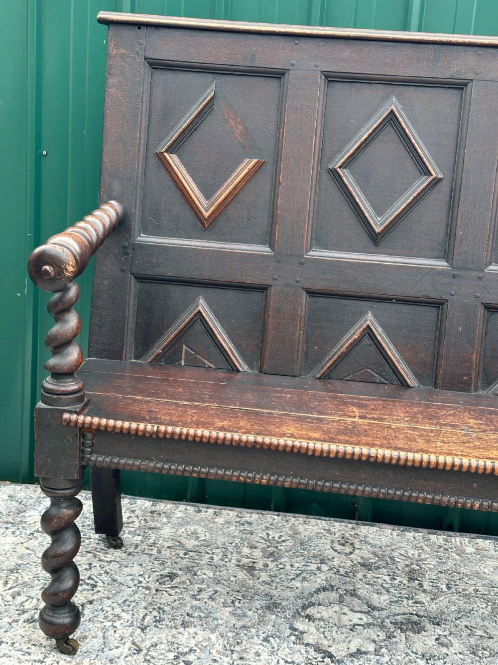 A dark oak 19th-century settle featuring a four-panel back with geometric diamond moulding, barley twist supports, and small brass castors. View II