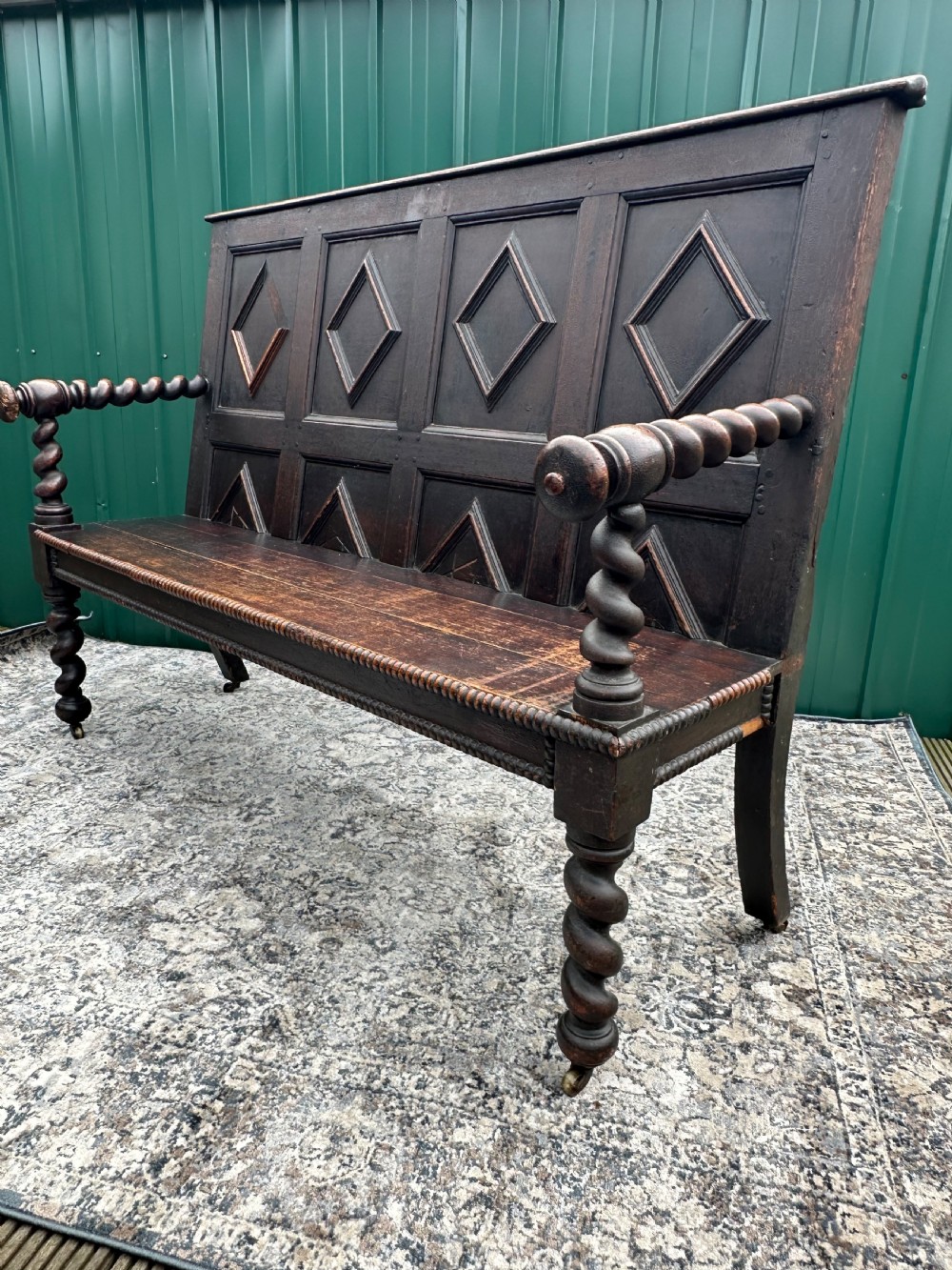 A dark oak 19th-century settle featuring a four-panel back with geometric diamond moulding, barley twist supports, and small brass castors. View V