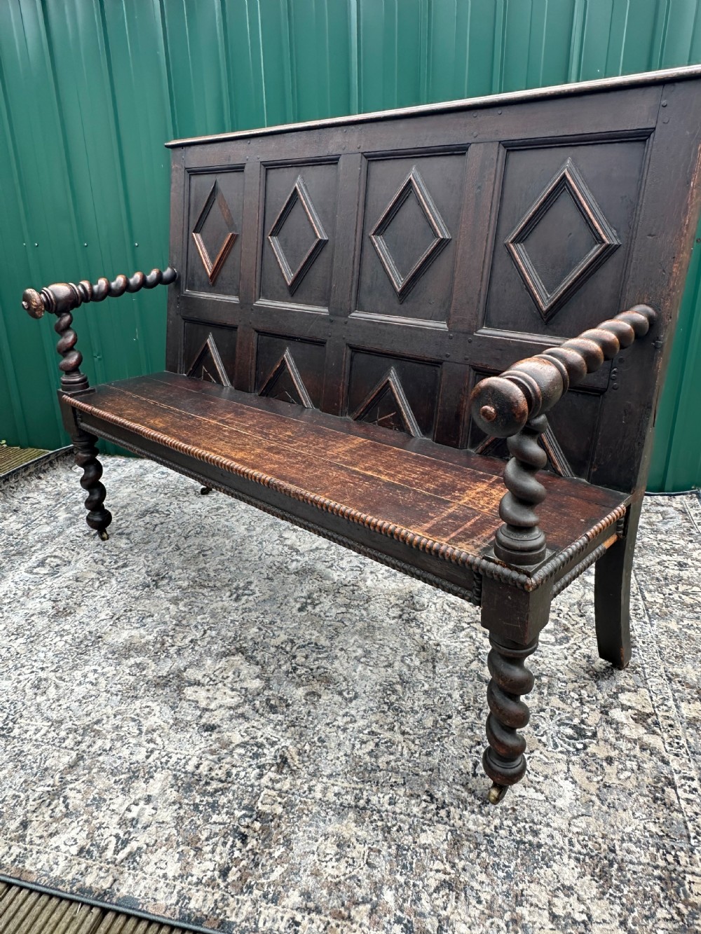 A dark oak 19th-century settle featuring a four-panel back with geometric diamond moulding, barley twist supports, and small brass castors. View XIII