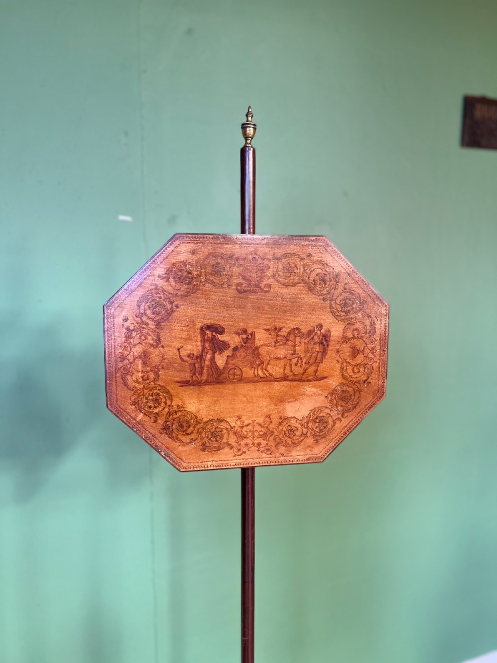 An antique Regency pole fire screen featuring an octagonal pokerwork panel supported on a turned wooden column with a plinth base resting on brass claw feet. View V