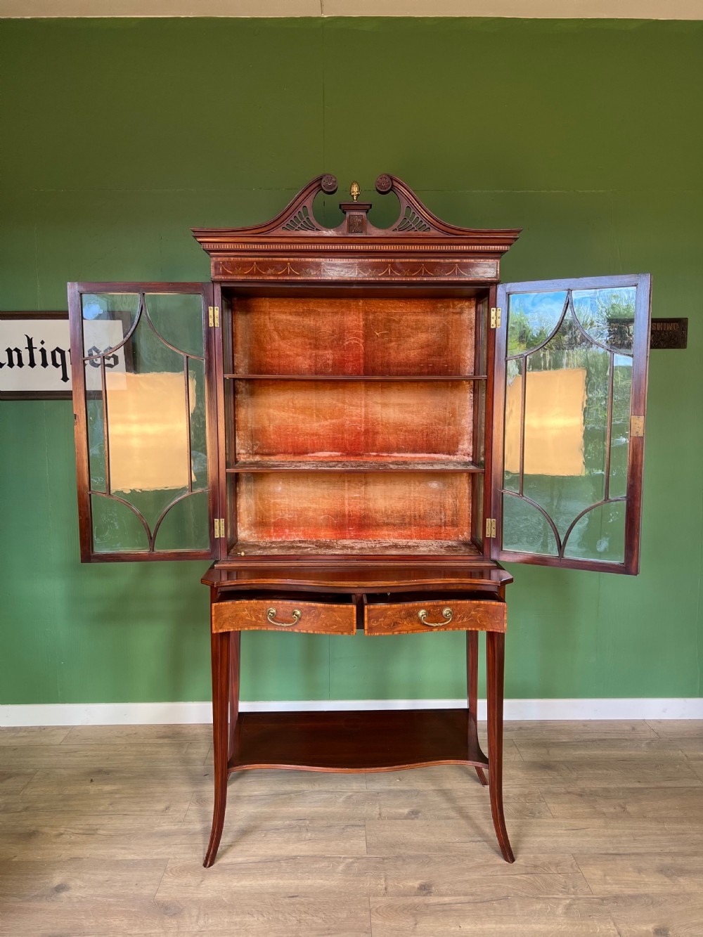 An Edwardian Sheraton Revival inlaid mahogany display cabinet featuring a swan-neck pediment, astragal glazed doors, drawers, and tapered legs. View II