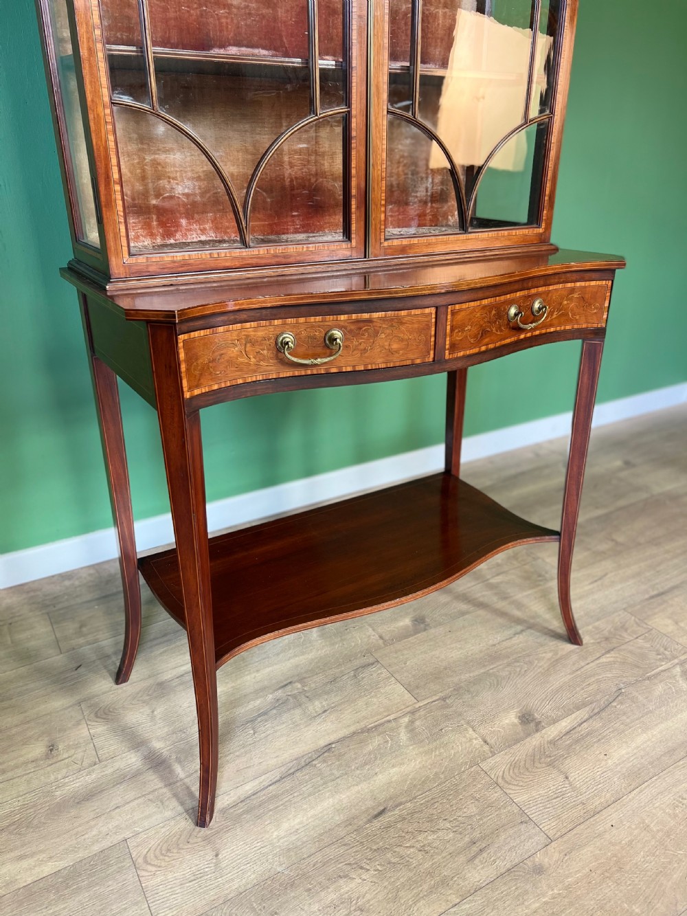 An Edwardian Sheraton Revival inlaid mahogany display cabinet featuring a swan-neck pediment, astragal glazed doors, drawers, and tapered legs. View IV