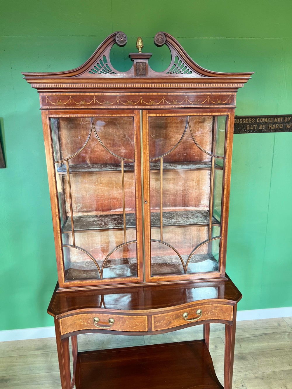 An Edwardian Sheraton Revival inlaid mahogany display cabinet featuring a swan-neck pediment, astragal glazed doors, drawers, and tapered legs. View V