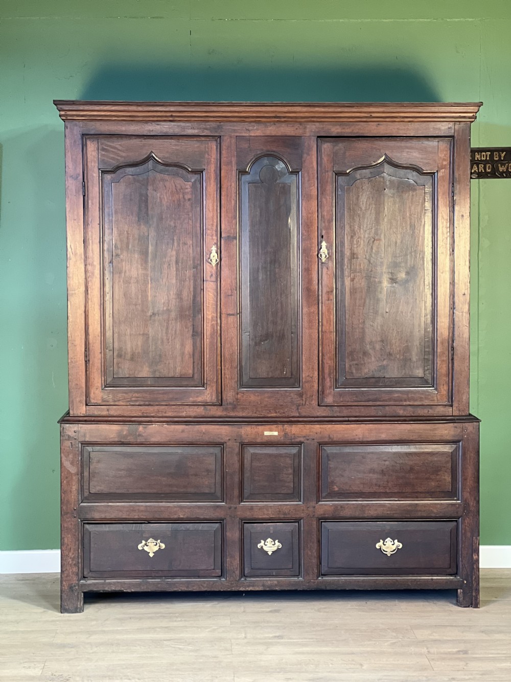 A mid-18th-century dark oak tack cupboard featuring arched panelled upper doors and a lower drawer section with brass handles.
