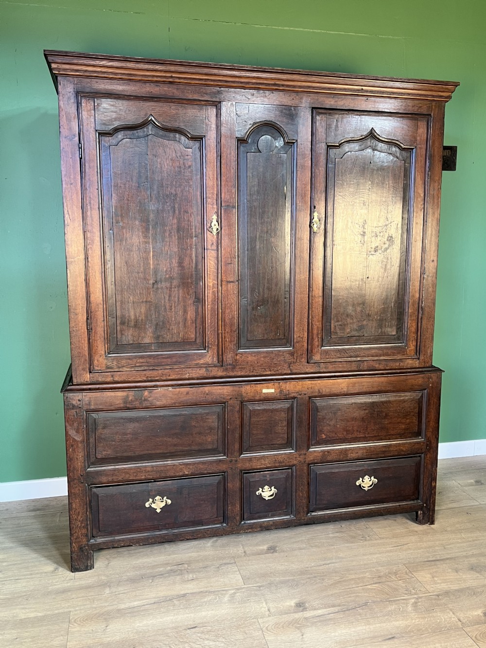 A mid-18th-century dark oak tack cupboard featuring arched panelled upper doors and a lower drawer section with brass handles. View II