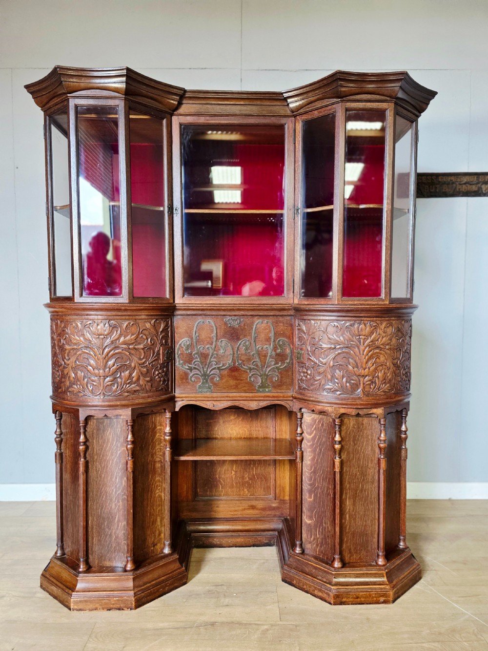 A late 19th-century carved oak cabinet desk with a red-lined glazed upper section, foliate carved lower panels, and brass strap hinges.