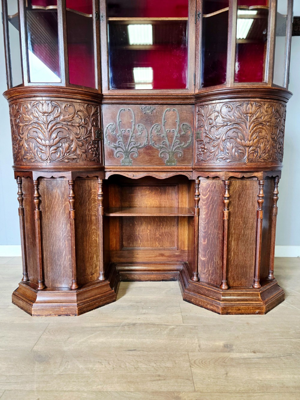 A late 19th-century carved oak cabinet desk with a red-lined glazed upper section, foliate carved lower panels, and brass strap hinges. View III