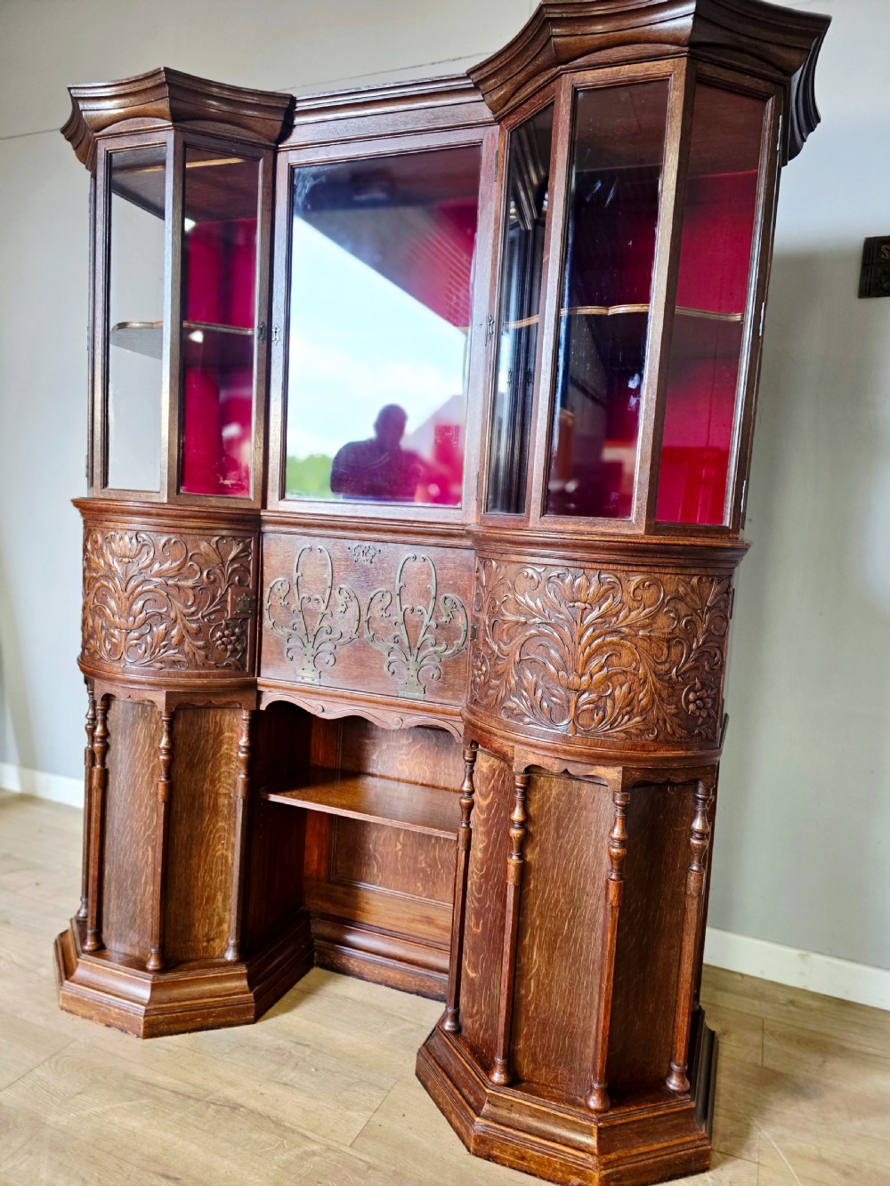 A late 19th-century carved oak cabinet desk with a red-lined glazed upper section, foliate carved lower panels, and brass strap hinges. View IV