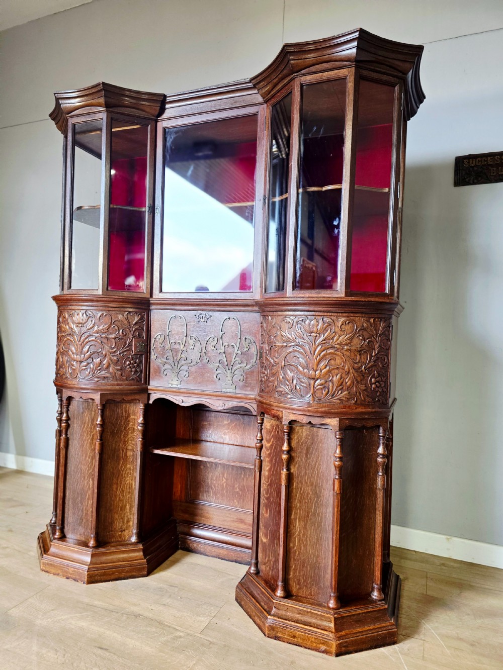 A late 19th-century carved oak cabinet desk with a red-lined glazed upper section, foliate carved lower panels, and brass strap hinges. View V