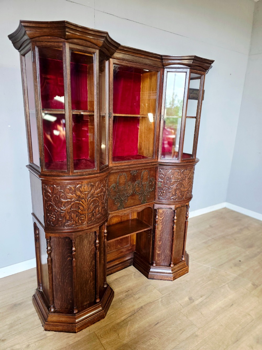 A late 19th-century carved oak cabinet desk with a red-lined glazed upper section, foliate carved lower panels, and brass strap hinges. View VI