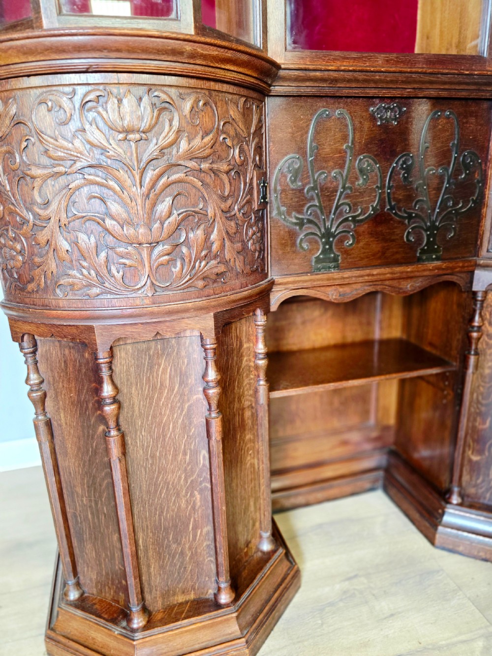 A late 19th-century carved oak cabinet desk with a red-lined glazed upper section, foliate carved lower panels, and brass strap hinges. View X