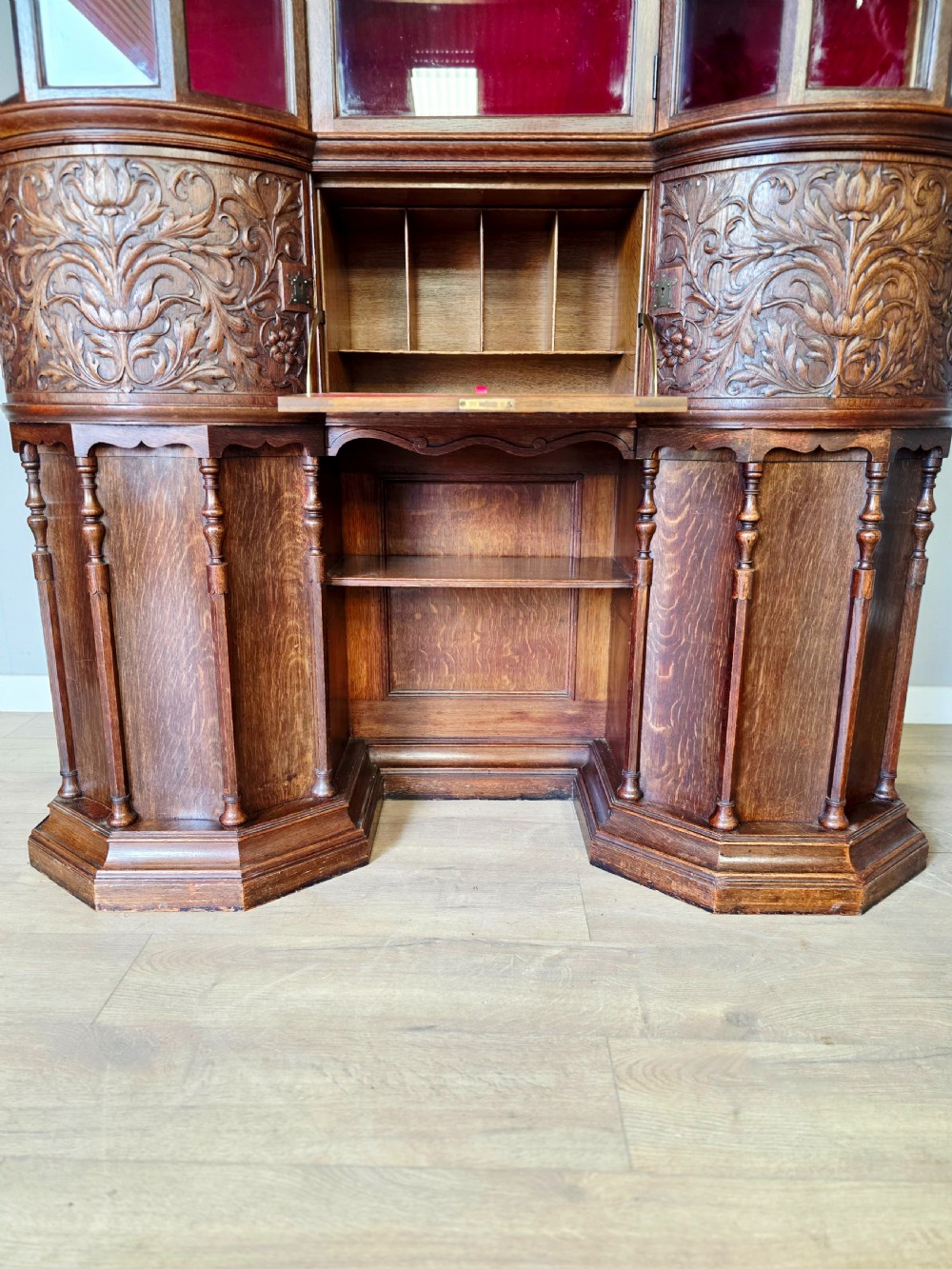 A late 19th-century carved oak cabinet desk with a red-lined glazed upper section, foliate carved lower panels, and brass strap hinges. View XI