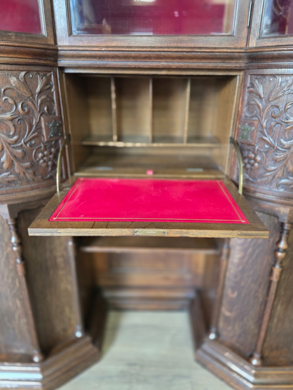 A late 19th-century carved oak cabinet desk with a red-lined glazed upper section, foliate carved lower panels, and brass strap hinges. View XII