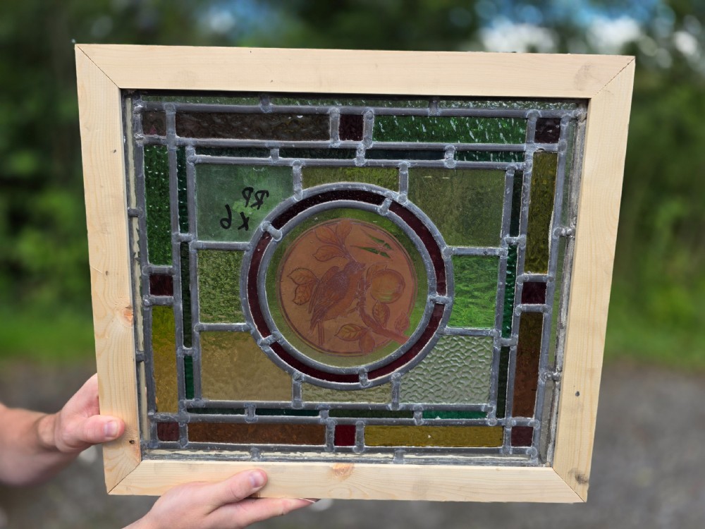 A square leaded glass panel featuring a central roundel depicting a bird on a branch surrounded by geometric green, amber, and red glass panes set in a light timber frame.