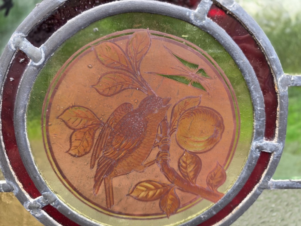 A square leaded glass panel featuring a central roundel depicting a bird on a branch surrounded by geometric green, amber, and red glass panes set in a light timber frame. View II