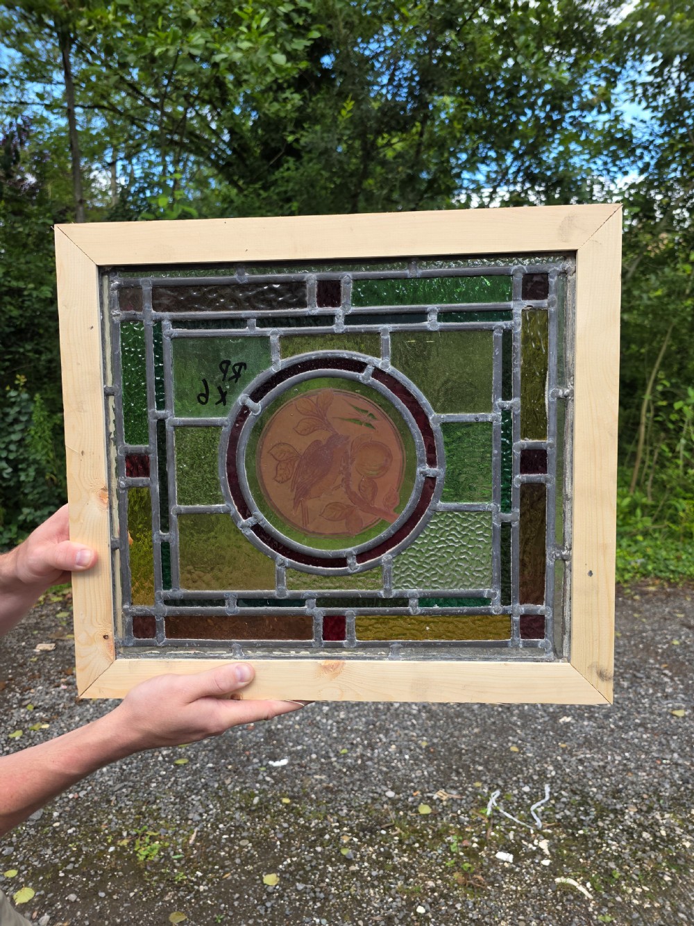 A square leaded glass panel featuring a central roundel depicting a bird on a branch surrounded by geometric green, amber, and red glass panes set in a light timber frame. View III