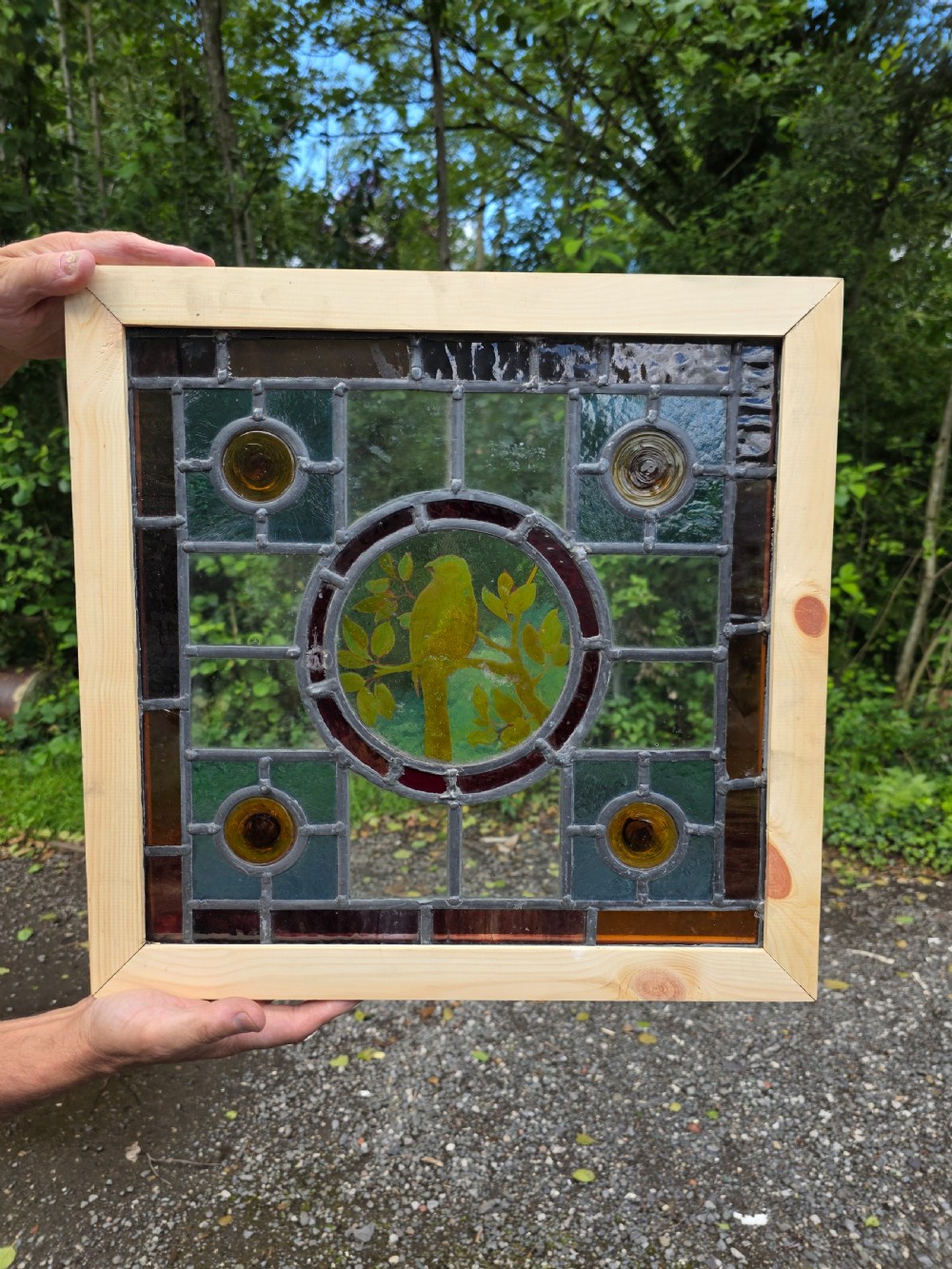 A square leaded stained glass panel featuring a central circular painted yellow bird on a branch, four amber bullseye roundels, and a light wood frame.