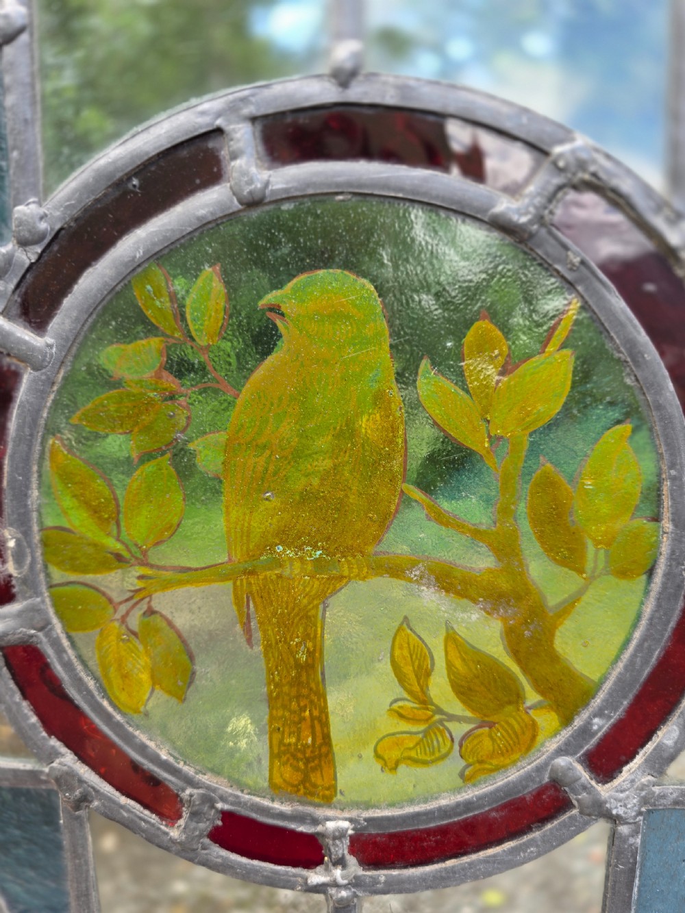 A square leaded stained glass panel featuring a central circular painted yellow bird on a branch, four amber bullseye roundels, and a light wood frame. View III